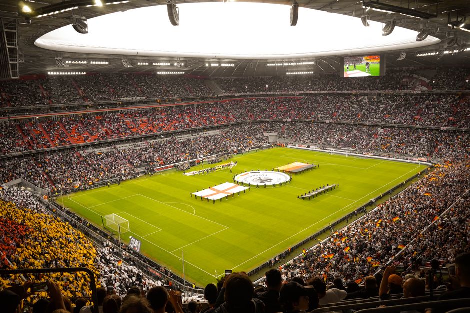 Inside a large football stadium during a match day, the pitch is illuminated with vibrant green grass and marked with white lines for gameplay. The seating areas are filled with spectators, many wearing white, black, and orange clothing, creating a lively atmosphere. At the center of the field, there are several large white and red sheets of fabric, possibly being used as part of pre-match preparations or ceremony, with some individuals standing nearby. Adjacent to these, a circular object is visible, possibly a large flag or banner, with people gathered around the edges. The stadium's roof features bright overhead lighting and a large, bright circular skylight allowing natural light to filter in. On one side, a large screen displays live footage of the match. The scene captures the environment of a professionally organized event, with the focus on the field where a home relocation of furniture and equipment for the match would be managed during non-game times, with Fulham Removals potentially involved in logistics or support for the event planning.