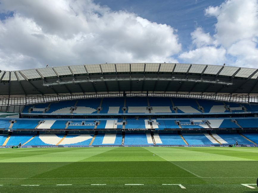 Inside a large football stadium during a match day, the pitch is illuminated with vibrant green grass and marked with white lines for gameplay. The seating areas are filled with spectators, many wearing white, black, and orange clothing, creating a lively atmosphere. At the center of the field, there are several large white and red sheets of fabric, possibly being used as part of pre-match preparations or ceremony, with some individuals standing nearby. Adjacent to these, a circular object is visible, possibly a large flag or banner, with people gathered around the edges. The stadium's roof features bright overhead lighting and a large, bright circular skylight allowing natural light to filter in. On one side, a large screen displays live footage of the match. The scene captures the environment of a professionally organized event, with the focus on the field where a home relocation of furniture and equipment for the match would be managed during non-game times, with Fulham Removals potentially involved in logistics or support for the event planning.