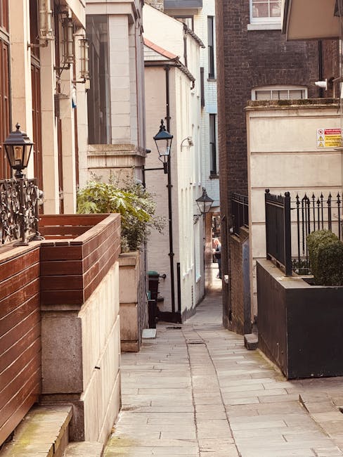 A narrow residential alleyway in Fulham with a paved pathway flanked by a red brick wall and a black metal fence on the right side, and a beige stone wall with a black street lamp on the left. The alley extends toward a distant street visible between closely spaced buildings with traditional brick and white facades. The scene is illuminated by natural daylight, highlighting the textures of the materials used in the construction of the buildings and fencing. This location, part of a house removal process, illustrates the constrained access often encountered in urban renovations, with potential space constraints for furniture transport and moving equipment. Fulham Removals regularly manages such narrow street moves, involving careful planning for loading and unloading furniture and boxes, often requiring packing materials like cardboard boxes and protective blankets for safe transport. The image captures the typical environment faced during home relocations in the Fulham area, emphasizing the importance of professional moving services in tight urban settings.