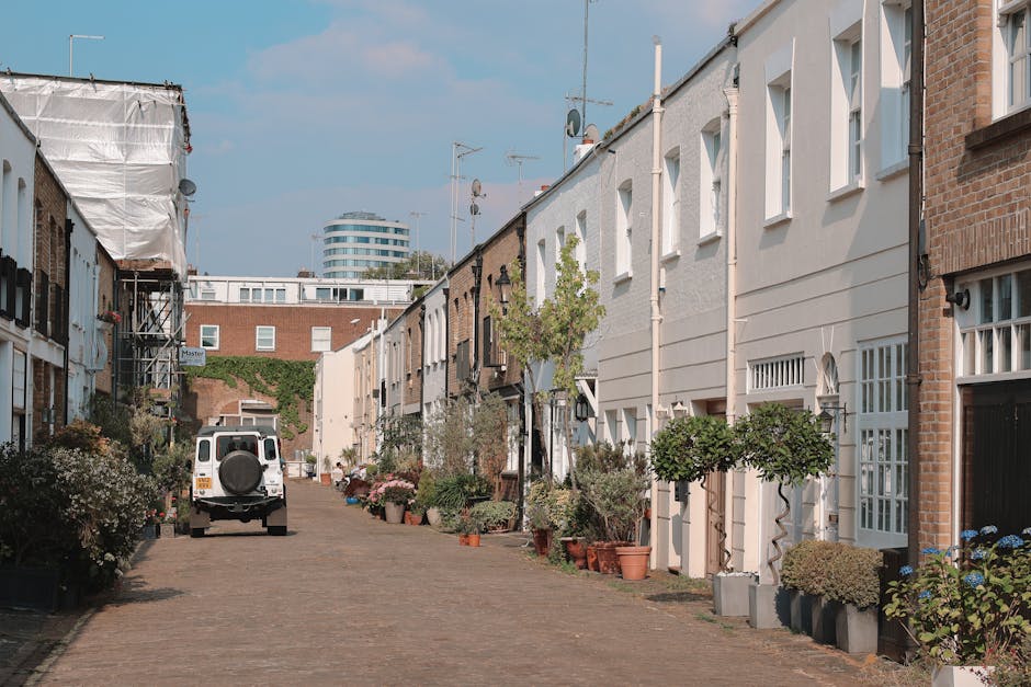 A narrow residential street with cobblestone paving, flanked by two- to three-storey terraced houses on either side. The houses have a mix of brick and painted exteriors, with white-framed windows and small front gardens filled with potted plants, small trees, and decorative shrubs. Some homes have outdoor lighting fixtures mounted near doorways. A white van with a spare tyre mounted on the rear is parked on the street, suggesting a home relocation or furniture transport process. Nearby, a few house fronts show signs of having scaffolding or protective sheeting, indicating ongoing refurbishment. The street appears to be in daylight with clear visibility, and a modern office building is visible in the background beyond the row of houses, emphasizing the urban setting. The scene captures the logistics of loading or unloading furniture and household items as part of a house move, consistent with the services offered by Fulham Removals, especially in complex narrow street environments.