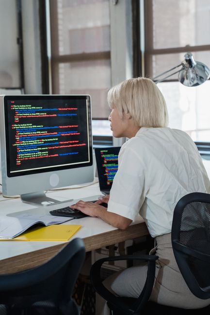 A woman with short blonde hair is seated at a desk inside a modern office space with large windows, working on a desktop computer displaying lines of colorful computer code. Next to her, a tablet also