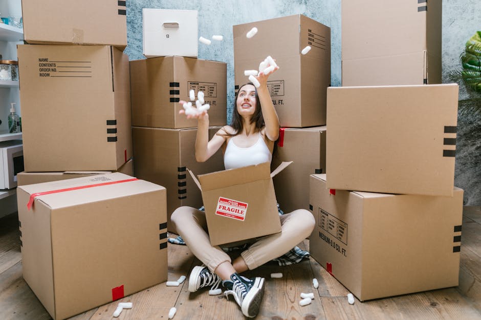 A woman sitting cross-legged on a wooden floor inside a home surrounded by numerous packed cardboard boxes of various sizes, some with labels and packing tape. She is wearing a white tank top, beige pants, and sneakers, and is smiling while tossing packing peanuts into the air. A partially open box in her lap contains packing materials, and more packing peanuts are falling around her. The background shows a textured wall, a potted plant on the right, and a kitchen area on the left. The scene captures the process of packing and preparing for a home relocation, with clear focus on the boxes, packing materials, and the woman engaged in the packing and moving process, illustrating a typical household move coordinated by Fulham Removals.