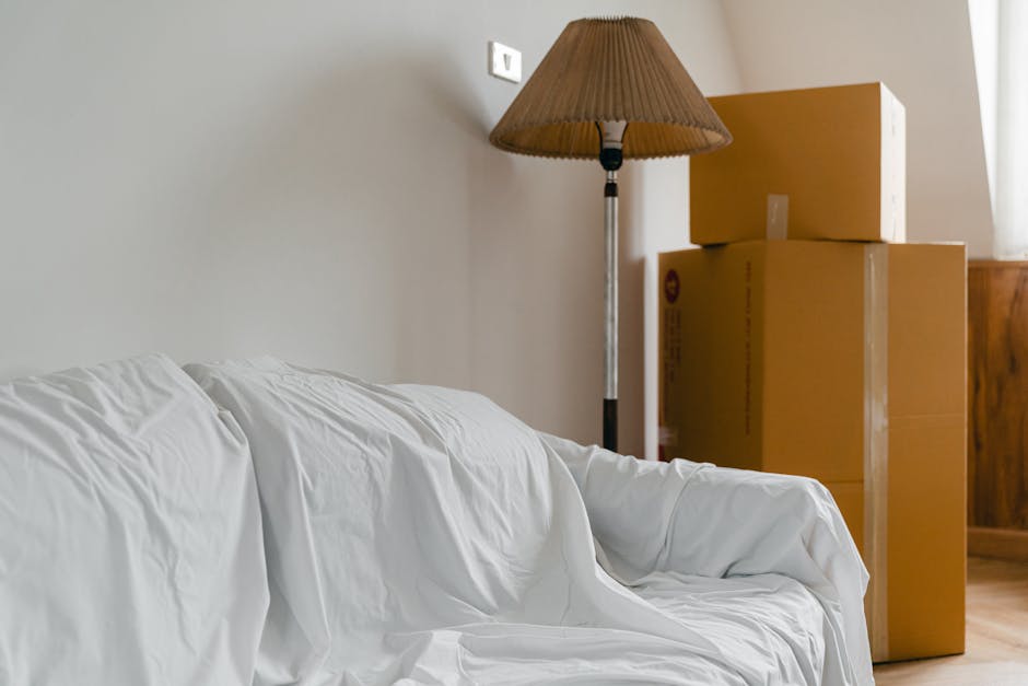 Interior scene showing a cardboard box placed on top of three other boxes, all stacked in a corner near a wooden sideboard and a window with white curtains. To the left, part of a sofa draped with a white sheet is visible, with the sheet slightly crumpled. A tall floor lamp with a tan shade stands adjacent to the boxes, providing ambient lighting to the room. The plain wall behind features a small white electrical outlet. The image depicts the process of packing or preparing items for a home relocation, consistent with furniture transport and moving logistics managed by Fulham Removals, illustrating elements of packing and stacking in a managed moving operation for a flat in Parsons Green, with natural lighting emphasizing the indoor environment.