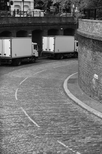 A black-and-white photograph depicts a curved cobblestone driveway in an urban area, with two medium-sized white moving vans parked side by side, partially visible in the background. The driveway is b